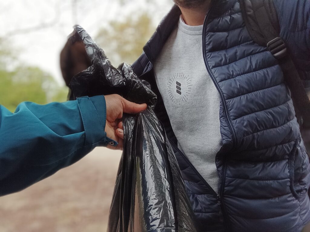 A close up of a man's chest as he holds up a bag of trash with another person just in the foreground holding up a second bag of trash they have collected from the ground.