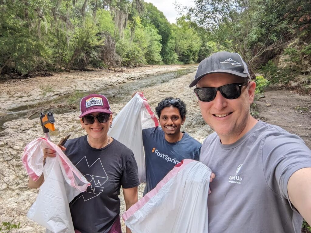 Three people hold up trash bags of garbage they collected from the waterway behind them.