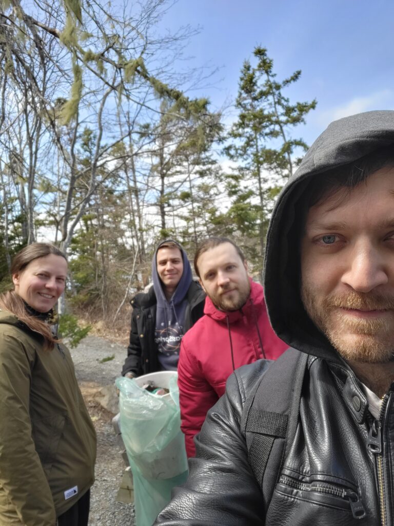 Four people in the woods are looking at the camera with one holding a bag of collected trash