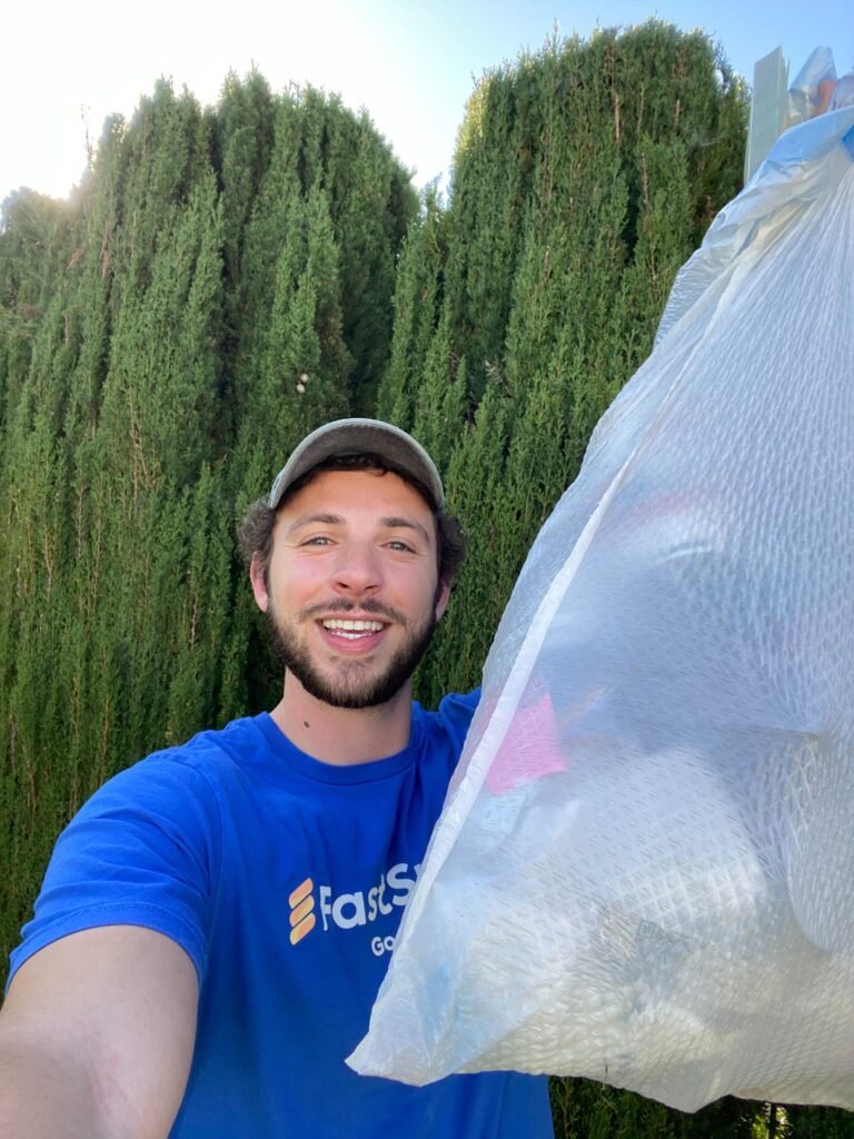 A man smiles at the camera while holding up a bag of trash in front of a row of tall arbovitae trees.