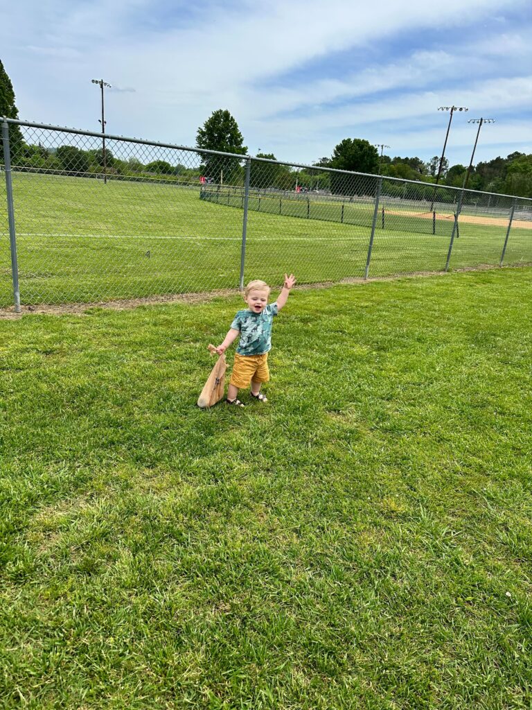 A small child holds a bag of trash in a field and is waving at the camera.