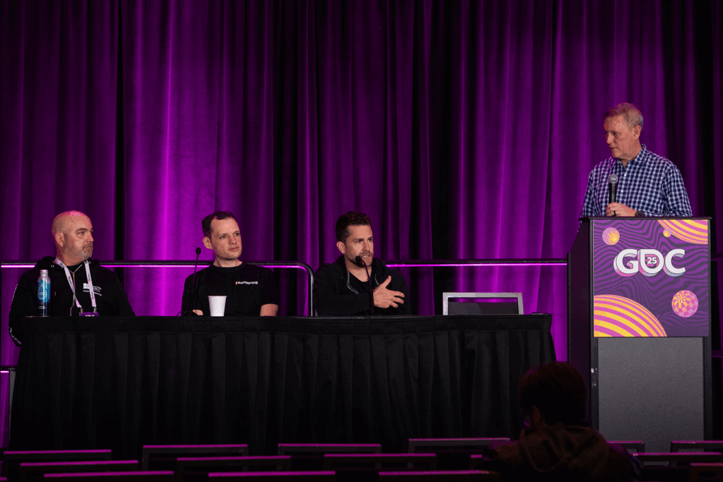 Chip sits between other panelists and David stands to emcee, all in front of a deep purple curtain.