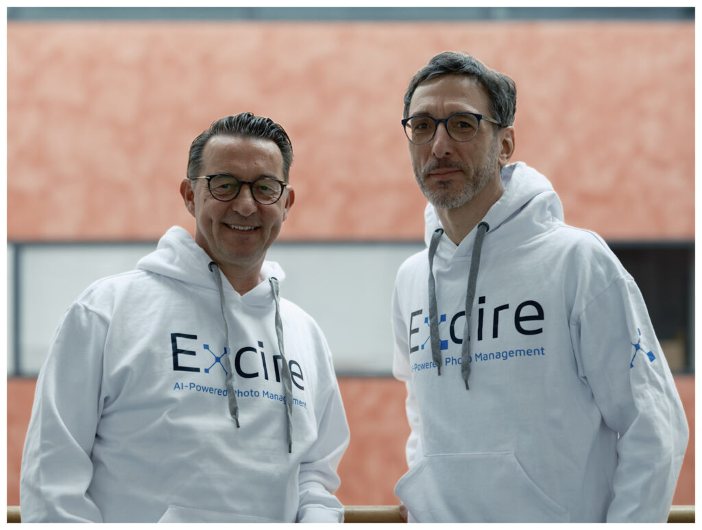 Mathias Martinetz and Thomas Käster wearing white Excire sweatshirts in front of a light brick wall.