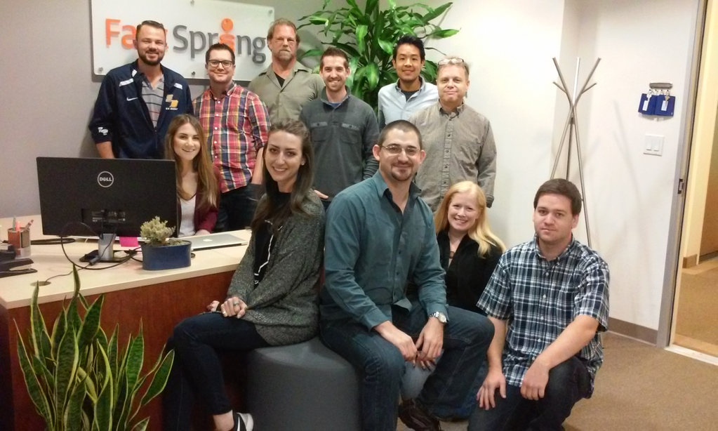 11 FastSpring employees stand or sit next to a desk in an office.