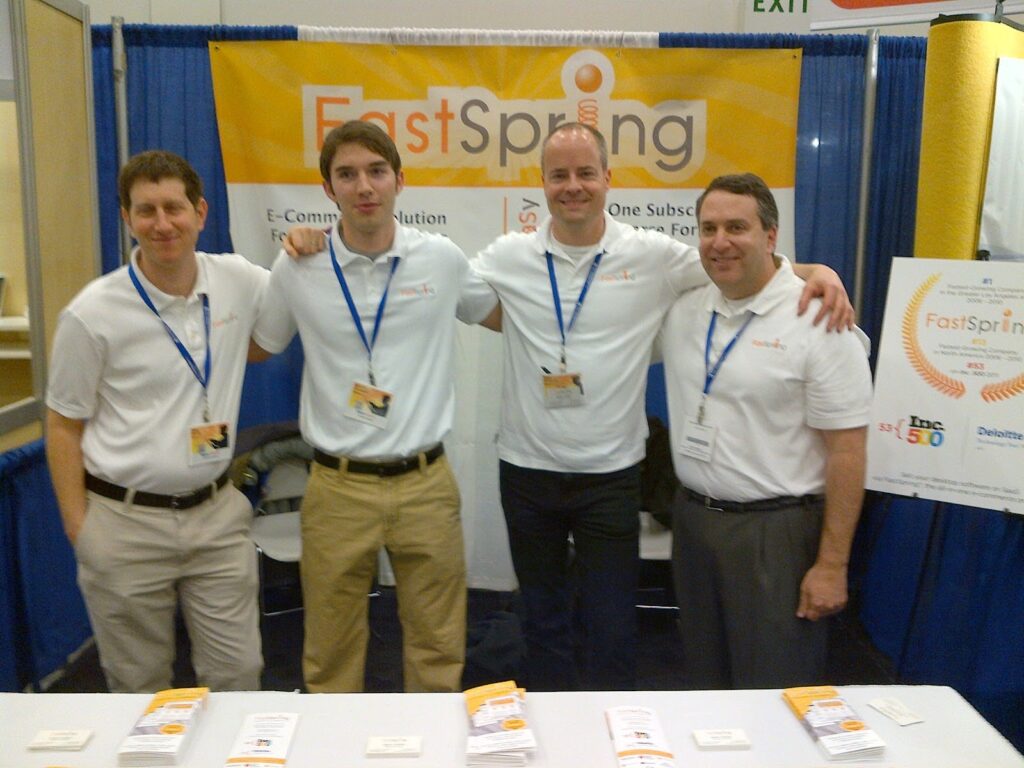 Four men stand in a trade show booth in front of a yellow and blue backdrop.