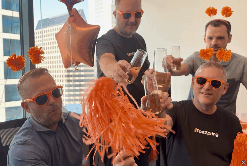 Four men wearing orange sunglasses and orange pompom headbands raise champagne glasses and an orange pompom for a toast.