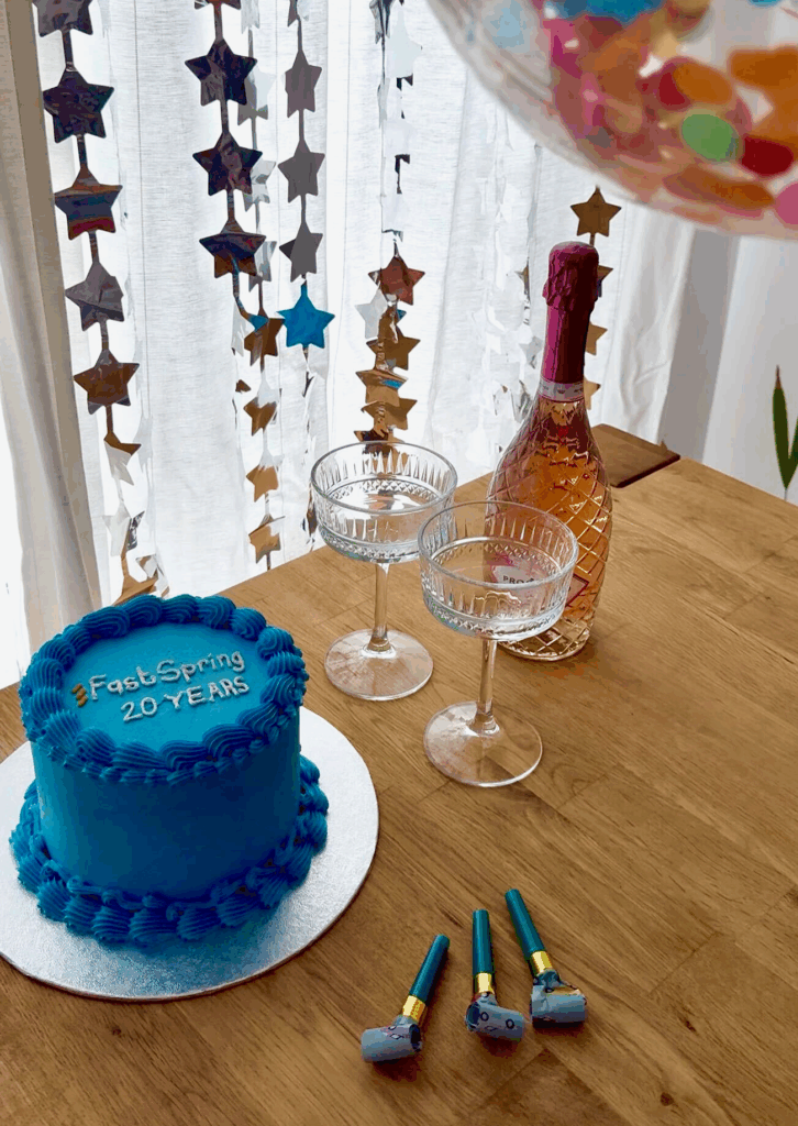 A blue cake site next to two coupe glasses and a bottle of rose wine, all sourrounded by a confetti balloon, star streamers, and party horns.