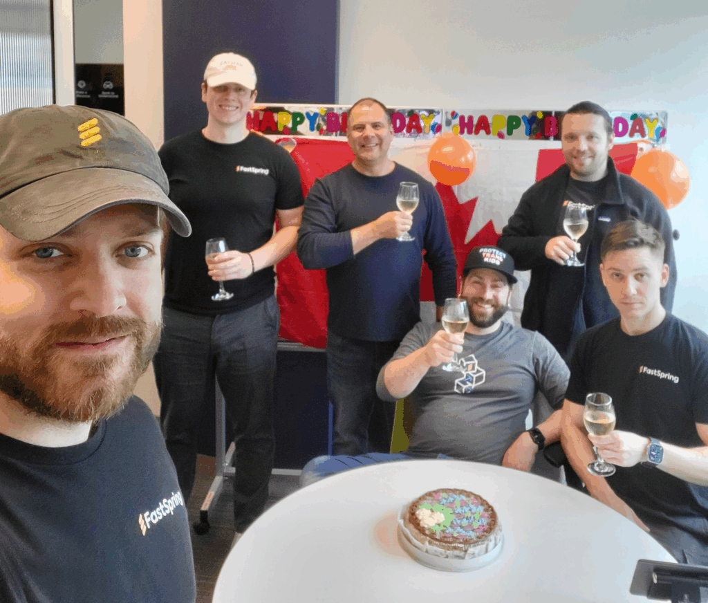 A man in the foreground takes a selfie with five coworkers in the background raising wine glasses in front of a Canadian flag and behind a Kyiv cake sitting on the table.