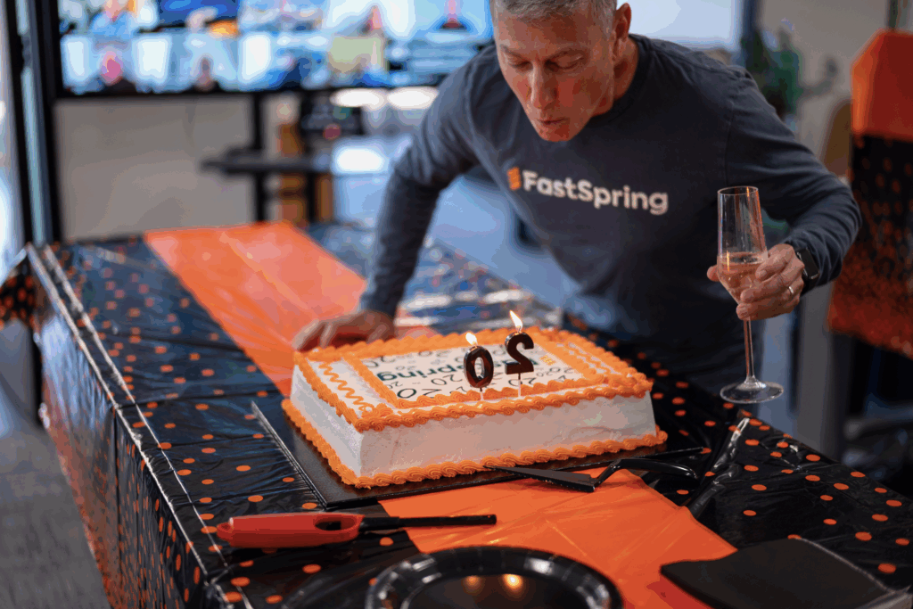 FastSpring CEO David Nachman leans over an orange and white cake with black 2-0 shaped candles to blow them out.