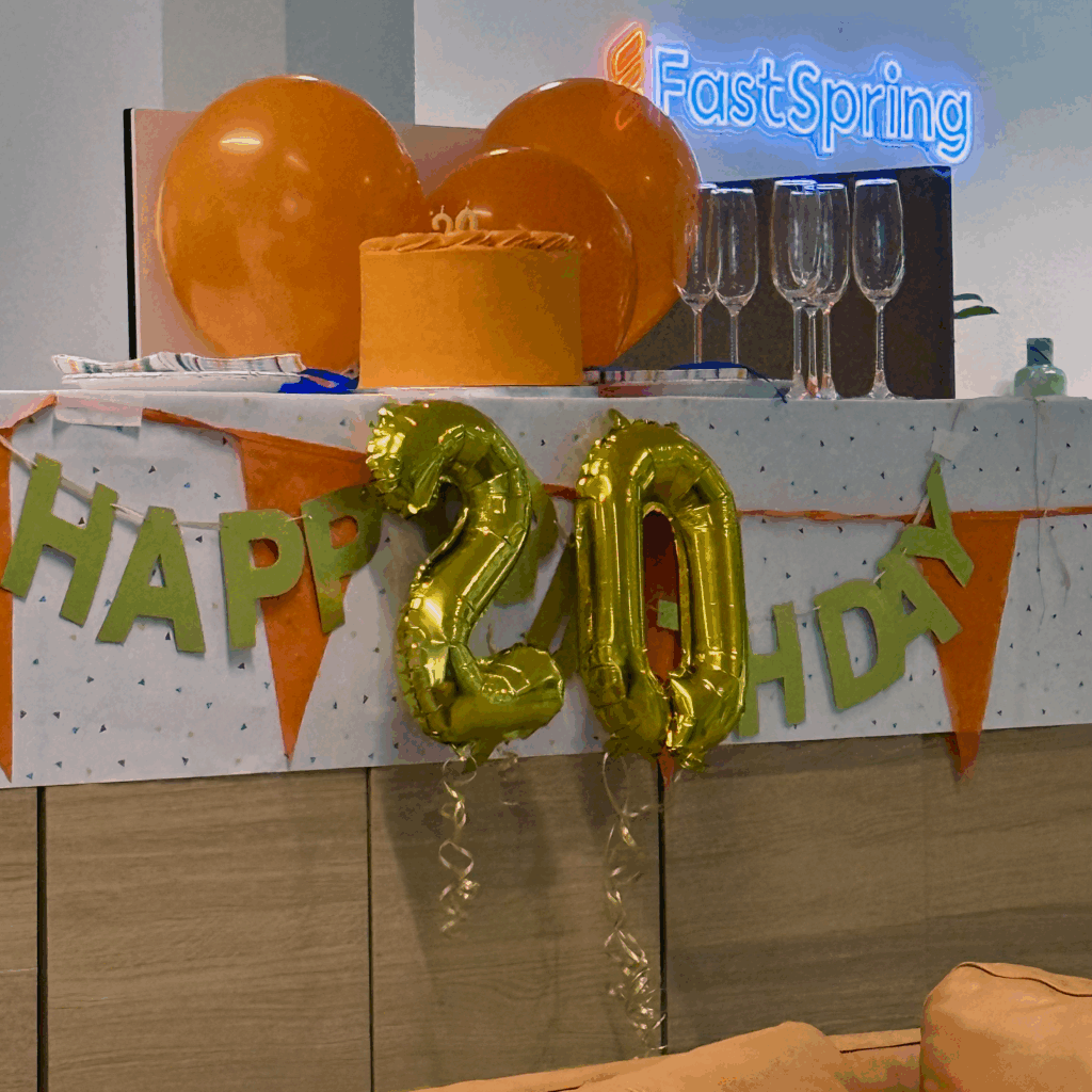 Orange latex balloons and an orange cake on top of a divider wall with metallic 2-0 balloons and a metallic Happy Birthday sign hanging below.