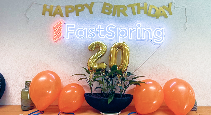 A metallic gold happy birthday sign hanging on a wall above a FastSpring logo neon sign, with gold metallic 2-0 balloons and orange latex balloons on the counter below.