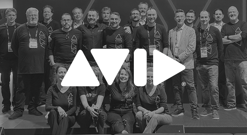 A black and white photo of a large group of people wearing black matching shirts and standing or sitting on a stage, with a white Avid logo superimposed on top.